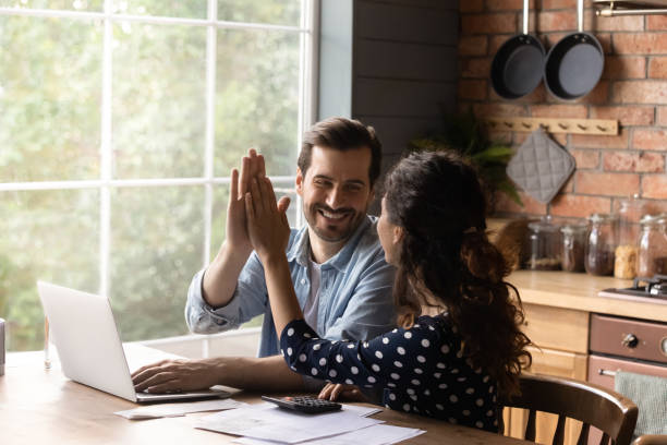 Happy young family couple giving high five to each other, finishing managing household budget, celebrating successful investment, making all payments online in e-banking computer application.